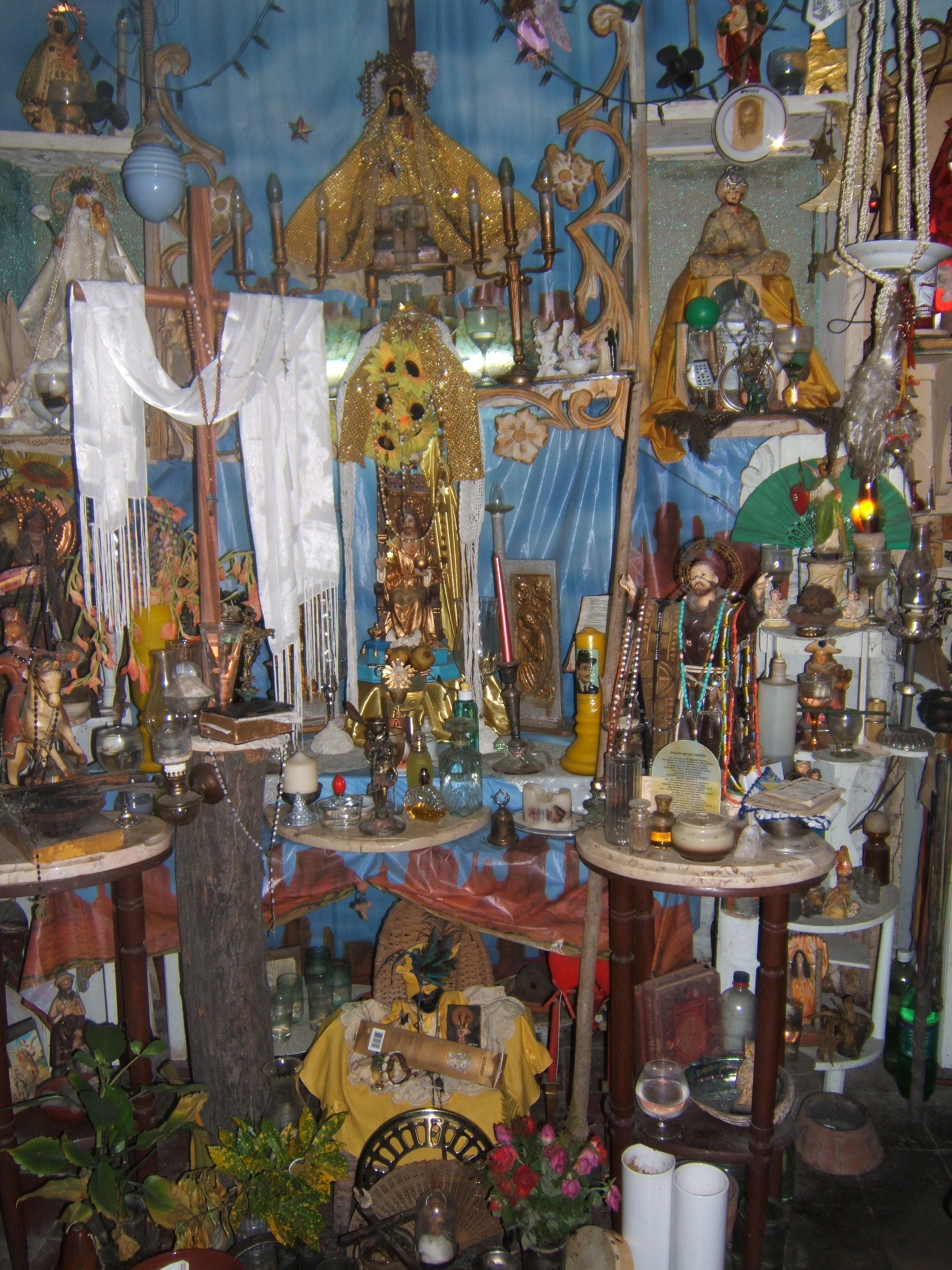 Figure 1. A view of the central altar in the altar room of Pedro, a Cuban religious practitioner from the doorway. The altar’s verticality is shown in multiple representations and doubling of the Virgin of Caridad del Cobre (in gold at apex) and the African spirit Josefina, identified with the oricha Ochún (in gold on floor). They are linked by a golden axis of other figures and adornments and surrounded by a host of other figures and offerings. Santiago de Cuba, August 2008. Photograph by author.