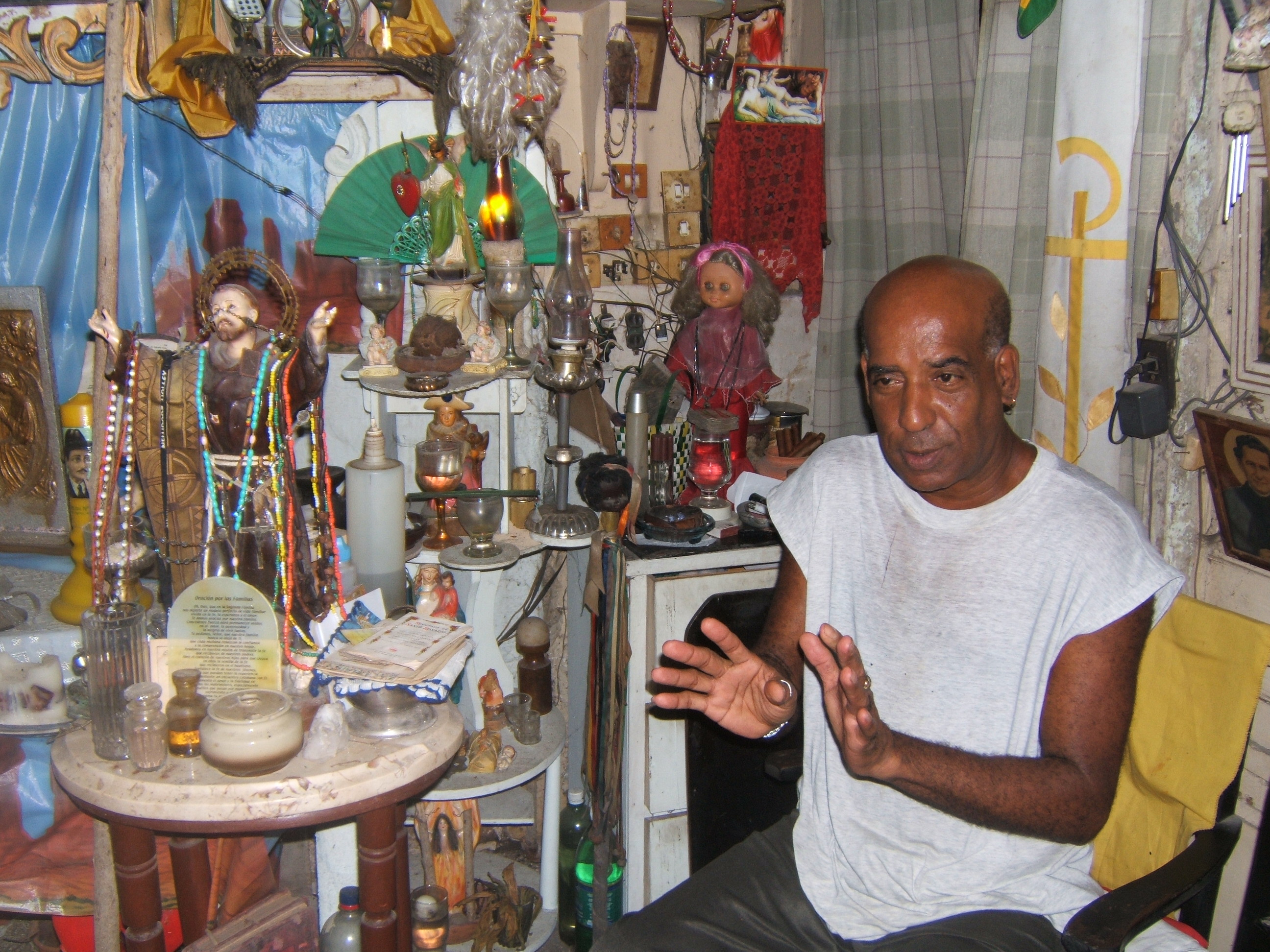 Figure 5. Pedro sits in front of a doll representing his Gypsy spirit (doll in pink), explaining something to the author. Next to him (left), a plaster figurine of San Francisco is draped with beaded necklaces (some representing orichas). He uses the booklet of prayers on the table to initiate spiritual consultations, in which he reads cards to receive images from the spirits. Santiago de Cuba, August 2008. Photograph by author.